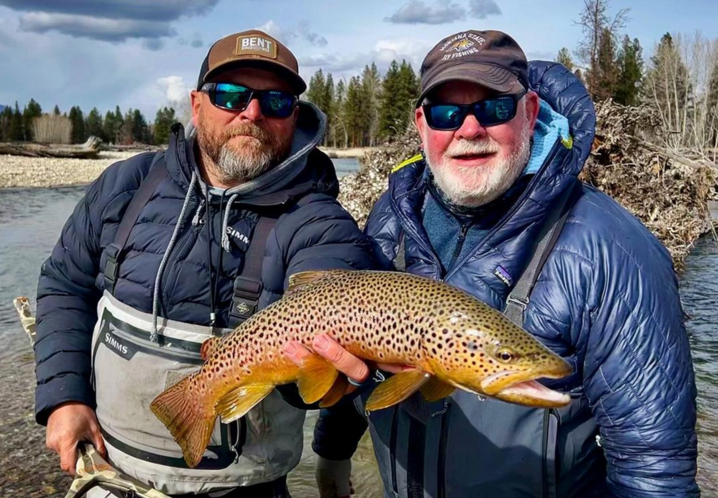 two men stand on a river while holding a brown trout caught while fly fishing